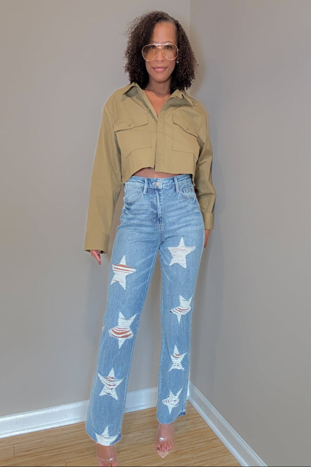 Woman wearing a beige jacket and jeans with star designs against a plain wall.