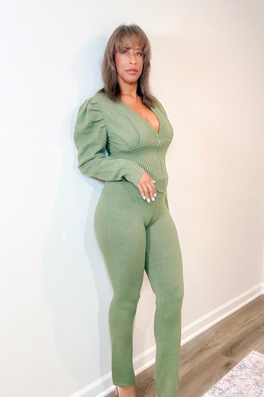 A woman modeling a sage green ribbed knit jumpsuit with long puff sleeves and a deep V-neckline, standing against a plain off-white wall.