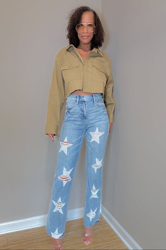 Woman wearing a beige jacket and jeans with star designs against a plain wall.
