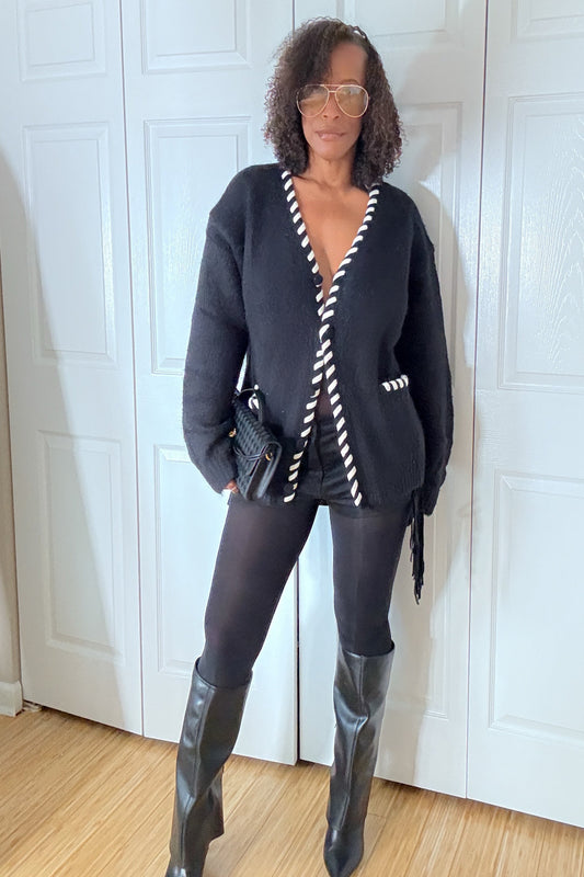 Woman wearing a black cardigan with white trim, black pants, and black knee-high boots in a room with white paneled walls.