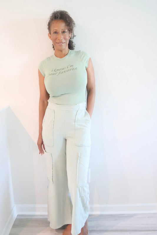 A woman stands indoors wearing a pale mint green short-sleeve t-shirt with black script text reading 'I know I'm your favorite' and off-white wide-leg pants.
